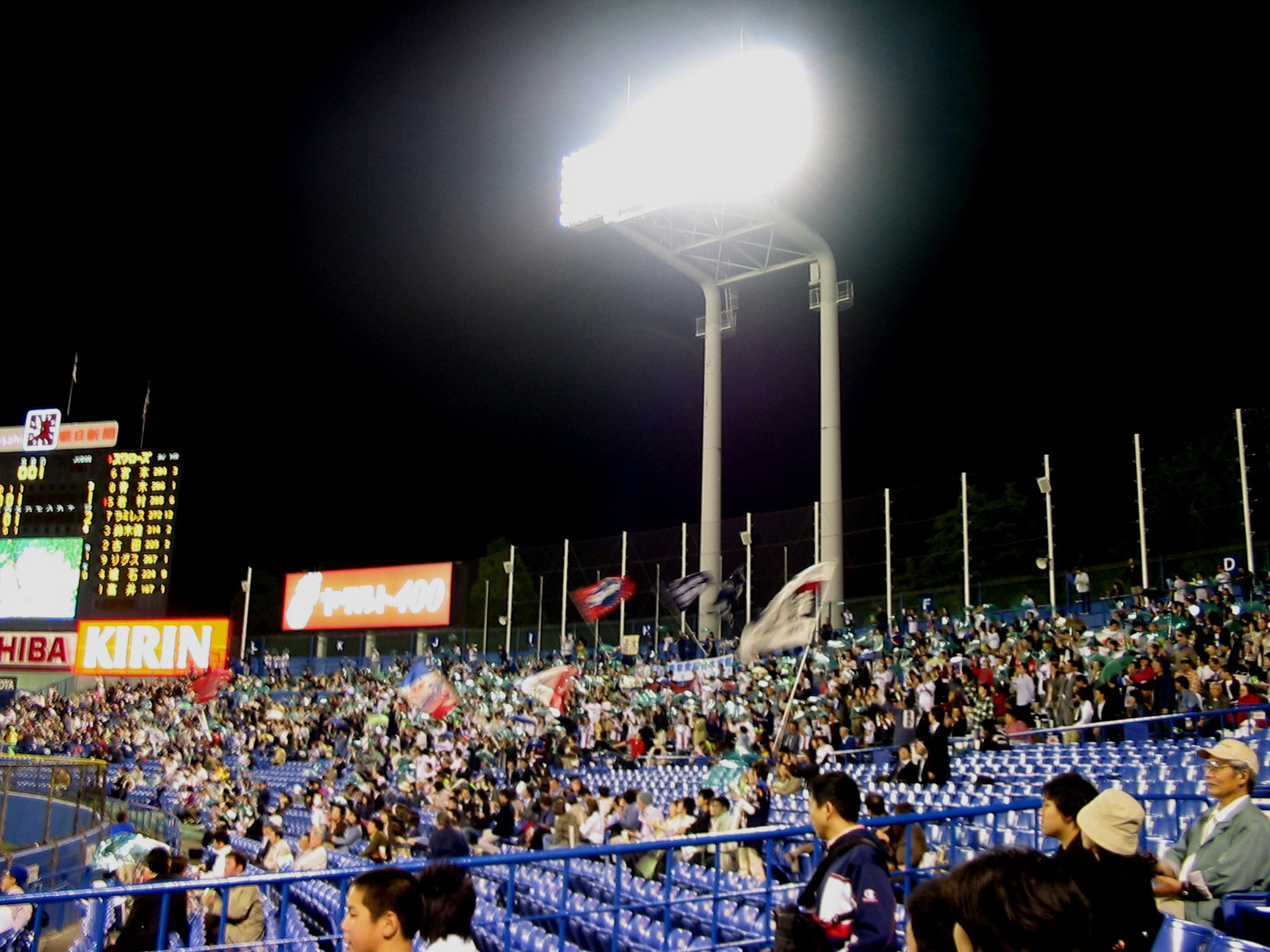view of baseball stadium from stands with bright lights and scoreboard